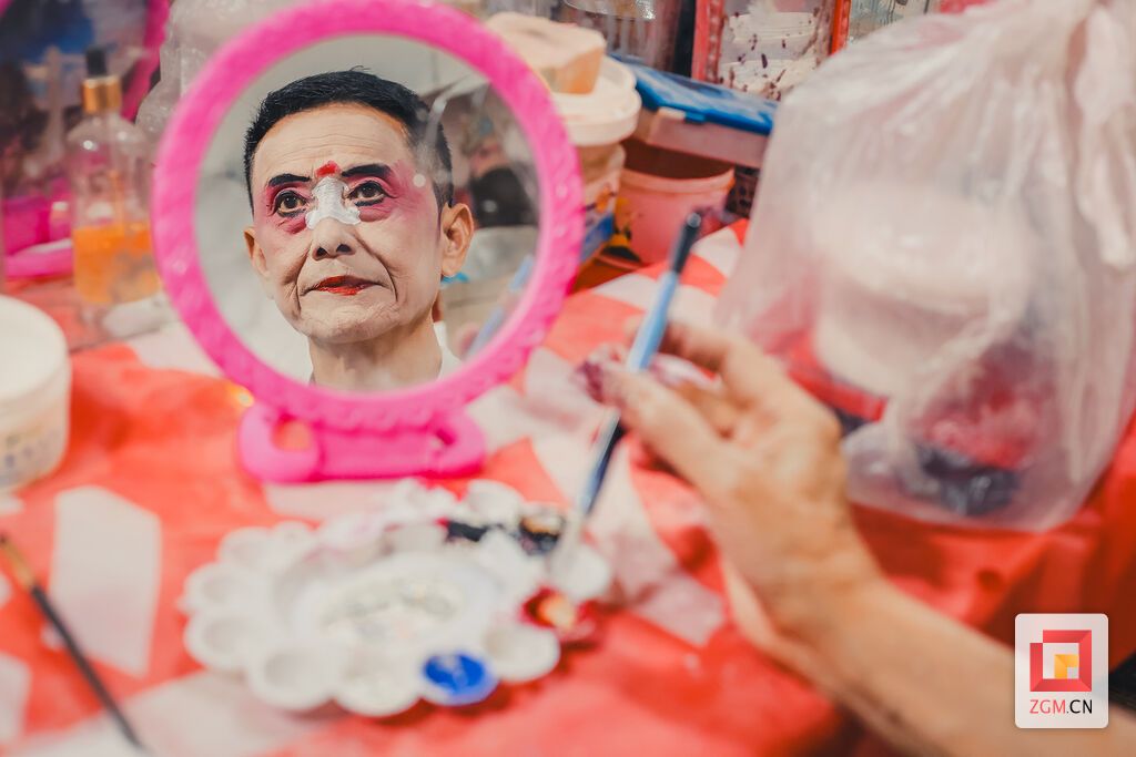 镜中映出他专注的眼神和半张已绘好的”脸谱,身旁摆满了各色油彩。.jpg